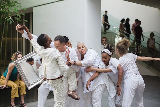 Performers dressed in white lean on each others backs in a line with the person in the front facing another person with one leg lifted in an angle and their extended hand touching the others. Back in the line one of the performers holds a mirror reflecting the leaning bodies.