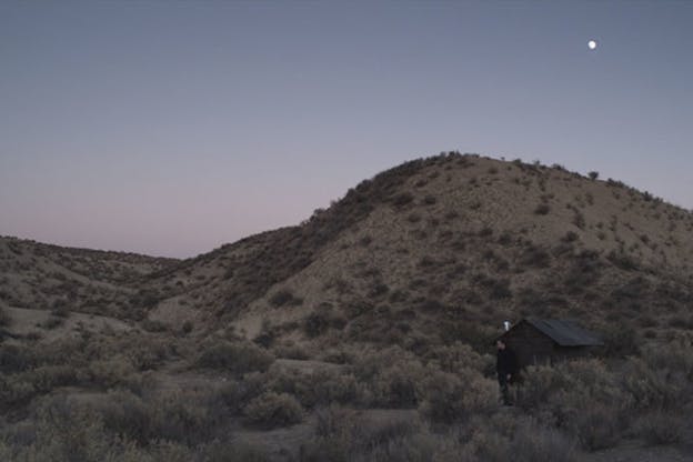 A video still of a person standing outside a small structure, in front of low hills dotted with small scrubs. The sky is a light purple with bits of pink near the bottom. In the upper right, the moon is visible. 