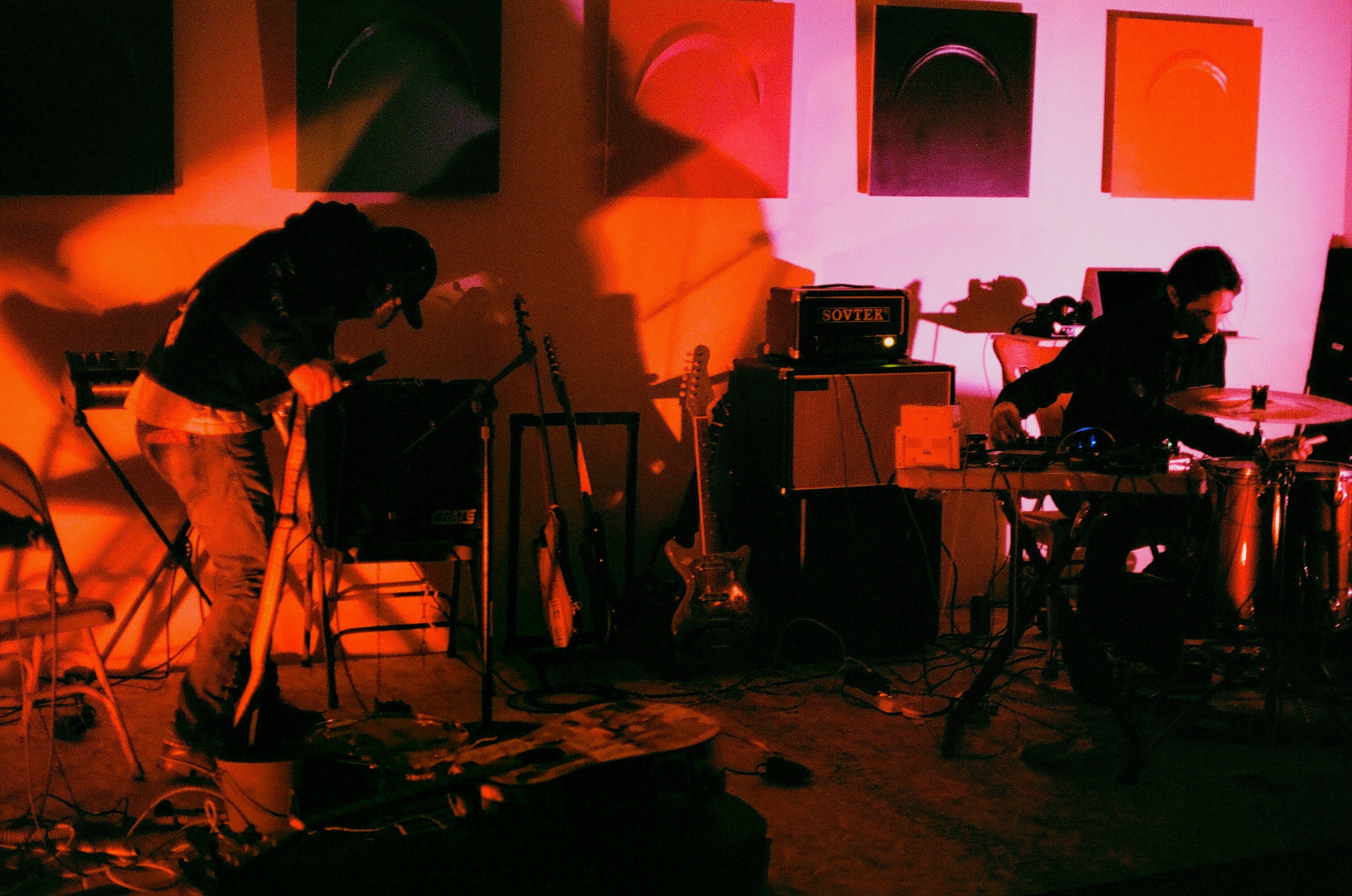 Awash in red light, Nathan Young and Mateo Galindo perform in a studio space scattered with instruments and equipment. Young stands on the left side of the frame, bent over with his face tilted away from the camera as he adjusts an amplifier. He is wearing jeans and a dark hoodie with a baseball cap. Opposite him, on the right side of the frame, Mateo Galindo sits behind a drum set with a small table of pedals and sound boards next to him. He gazes down at the drum before him with his right hand adjusting a soundboard.