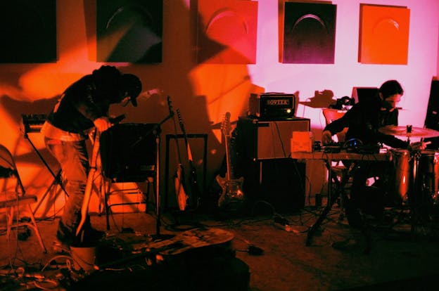 Awash in red light, Nathan Young and Mateo Galindo perform in a studio space scattered with instruments and equipment. Young stands on the left side of the frame, bent over with his face tilted away from the camera as he adjusts an amplifier. He is wearing jeans and a dark hoodie with a baseball cap. Opposite him, on the right side of the frame, Mateo Galindo sits behind a drum set with a small table of pedals and sound boards next to him. He gazes down at the drum before him with his right hand adjusting a soundboard.