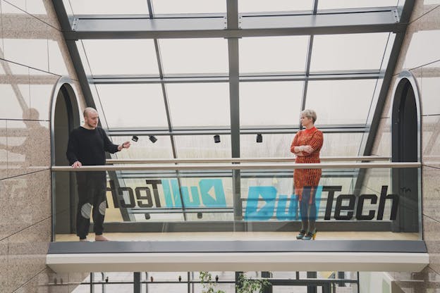 Two people stand far apart on a glass bridge with two tunnels on each end, inside a building.