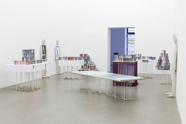 Installation view of tables stacked with multi-colored box-like objects with letters and geometric shapes that are increased in size and pasted to the chests of three dummies arranged against the walls. 