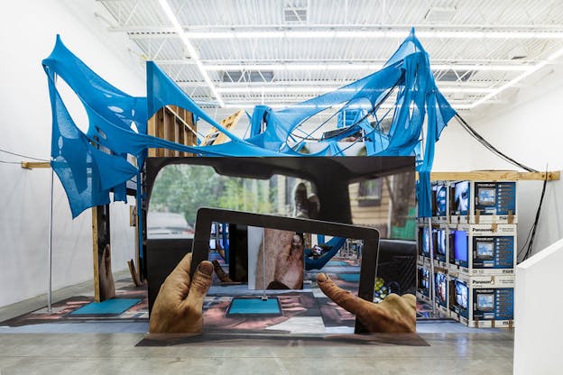 A large sculpture is within a white warehouse space. Several wooden frames are overlayed with stretched blue fabric with holes in it. In front of this, there is a large imafe of a person holding a screen which shows a part of a person's body in front of another scene. On the right, there are stacks of televisions projecting images.