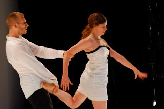 A performer in a white dress leans forward while another performer supports them by leaning backwards and holding the performer's left leg and right arm. 