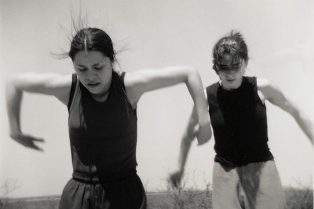 A black and white performance still of two performers jumping and wearing dark colored tank tops. The performer on the left looks downwards and has their arms lifted to ninety degree angles beside their body. The performer to the right looks downwards and has their arms raised unbent beside their body. In the background, there is a clear sky and a grass field. 