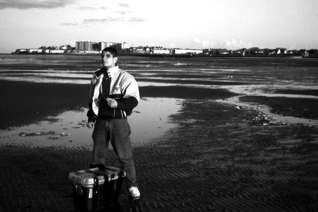 A black and white film still of a person wearing a jacket and jeans standing in front of a small plastic crate on a wide, expanse of wetland. In the disatnce behind them there is a town. The person looks up and to the left at something beyond the frame of the image.