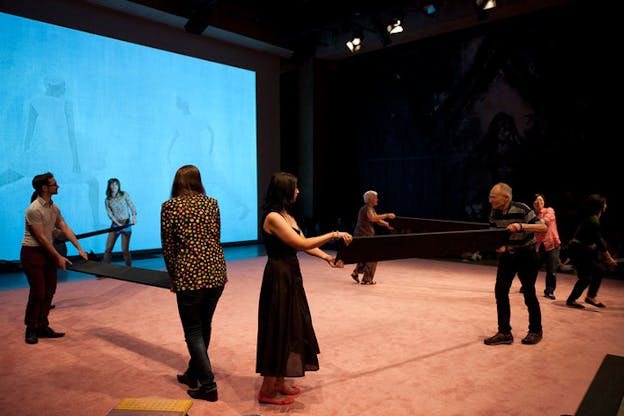 Persons hold the ends and center of two long 90-degree angle black planks on a reddish tan stage with a blue screen projecting images of persons lunging.