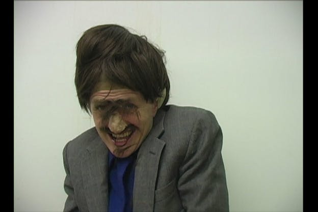 A video still of Ben Tor in front of a white wall, wearing a disorganized brown wig with scattered bits of hair on her face, a fake nose, and a grey suit jacket with a blue shirt. She hunches her shoulders and smiles at the camera. 
