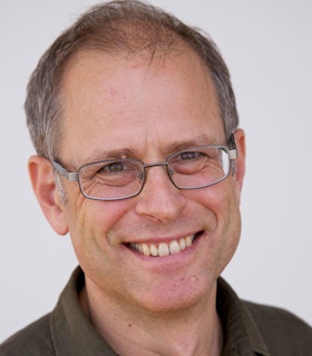 A portrait of Paul Kaiser smiling in front of white background. He wears a brown collared shirt, thin wire glasses, and has short grey hair.