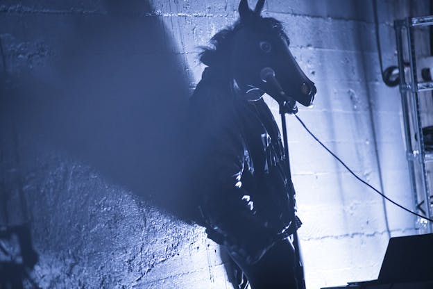 A figure wearing a horse head mask leans on a bare wall illuminated by white-gray light.