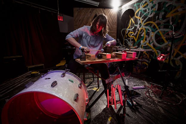 A sitted person looking down towards a stand supporting four identical wooden slim boxes separated by various lollipop shaped instuments, holds two sticks with round ends towards them. Cylinders hang from the legs of the stand and a drum stands in the front closest to the viewer. The room with cluttered with cables on the black floor has swirls of graffiti on the wall right to the figure.