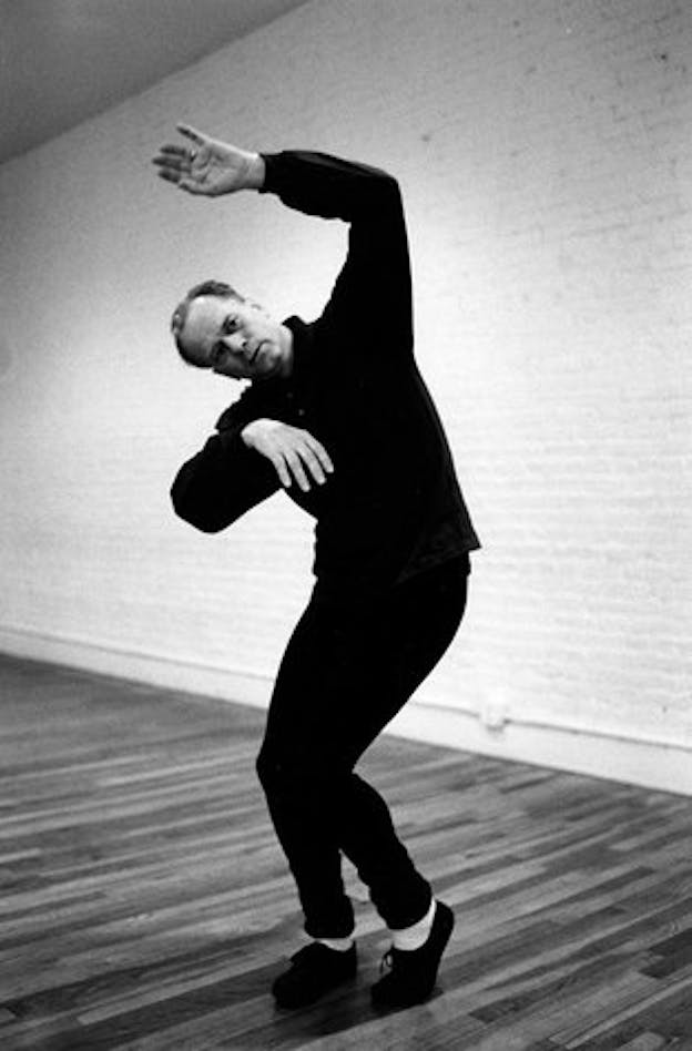 Black and white image of performer in a dancing movement. Their bended knees touch as their angled right hand reaches towards the front while the left angles on top of their head.