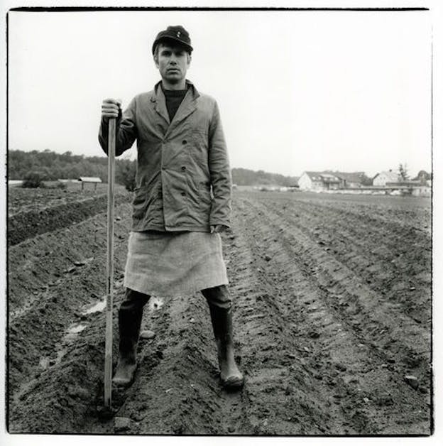 Black and white photograph of a person standing in a field, dressed in a coat, long shirt, plastic boots and a hat holding a hoe.   