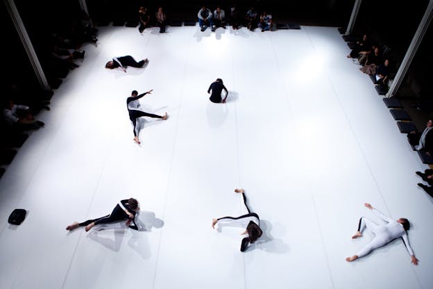 Eagle view of performers dressed in full body leotards, white in the front and black at the back, lay down in various poses on a white rectangle floorboard. Surrounding the stage an audience covered by the shadows.