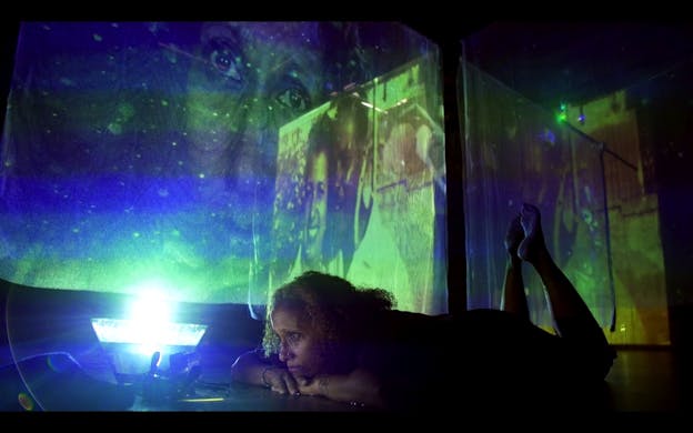 A performance still of nia love laying with her stomach on the ground and her feet up behind her, looking intently at a florescent blue lit cone in front of her. The setting is dark and love's body is encased in shadows. On the wall beside her there is a projection of an opaque image of two people standing close to each other and smiling with a translucent image of love's own face layered atop it. Blue, purple, and yellow light tints the scene.