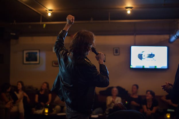 A person with their back to the viewer dressed in a black leather jacket and holding a microphone raises their hand up as an audience watches.