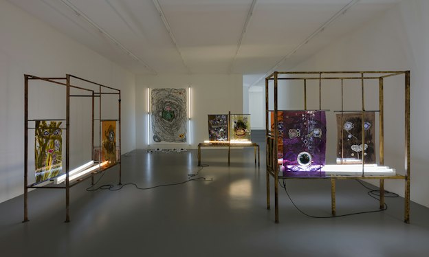 Steel structures support colorful abstract plexiglass with gem rocks glued on them depicting eyes and mouths. The art is illuminated from behind with long neon lights, their cables fallen on the floor in swirls connecting with each other.