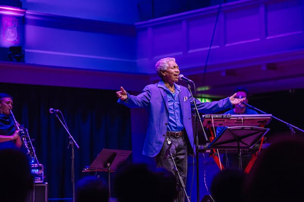 A performer wearing a purple dress jacket sings on a stage illuminated by purple lights. Instrumentalists sit behind him while in the front of the image closer to the viewer the back heads of the audience are blurred.