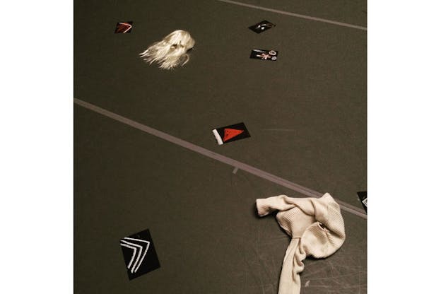 Film-grain photograph of a platinum wig and crumpled white sweater strewn beside black sheets drawn with white and red chalk across a tennis-green floor with two gray tape stripes.