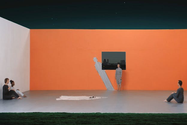 A person dressed entirely in stripes stands against a tangerine orange wall in front of a gray landscape image and a slanted cut-out striped figure. Persons in gray unitards sit against the adjacent walls and in the center of the floor is a flat white irregularly-shaped sheet. 