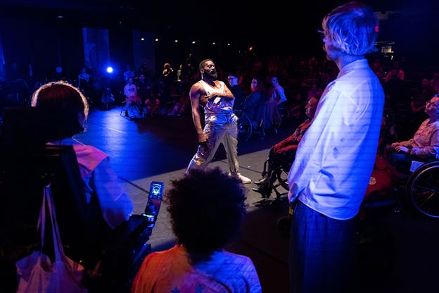 A performer stands on in the middle of a black box stage illuminated by purple and pink lights. The audience surrounding him stares.