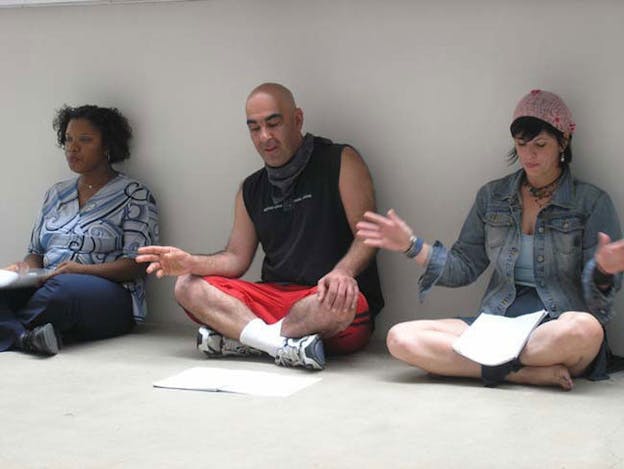 Three people sit cross-legged on a white floor while looking down at their individual stack of paper. 