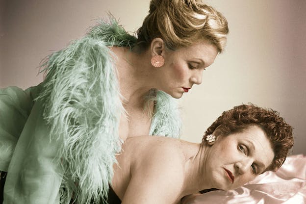 Two people are in front of a dusty rose colored background and laying on a similar colored silk. One person with short copper hair lays on their front on the rose silk and wears a low cut, strapless black top, and diamond earrings. They look back and up at the other person who lays on top of their back and wears a pastel green jacket with feathers around the neckline, pink earrings, red lipstick, and has their blonde hair pulled into an updo hairstyle.