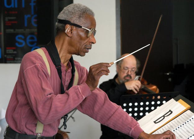 A person wearing glasses and looking at a music sheet. In their right hand they holds a baton, behind them blurred is a figure playing the violin. 