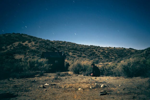 A performance still of van der Werve seated on the floor, with his legs crossed, besides a small structure, surrounded by low shrubs. Many small streaks of light are visible in the sky. 