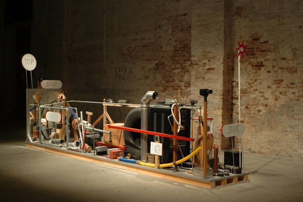 A low horizontally long platform with various pieces such as tubes, wheels and wood. In the front closest to the viewer, the shorter side holds a small sign with the word 