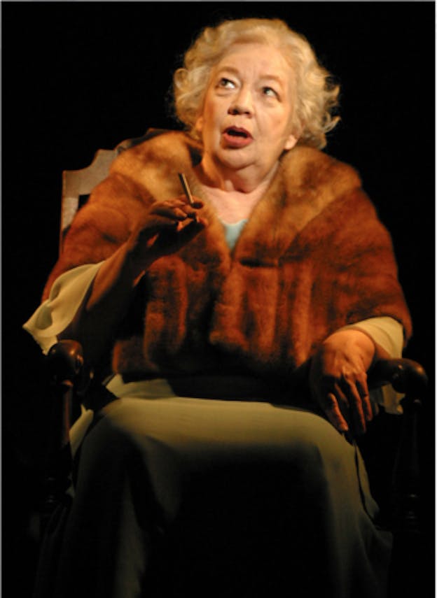 An image of Maleczech performing in a dark space, wearing a green dress, fur stole, and with short blonde curly hair. She looks upwards and opens her mouth, appearing to be mid-speech. In her right hand, she holds a cigarette.