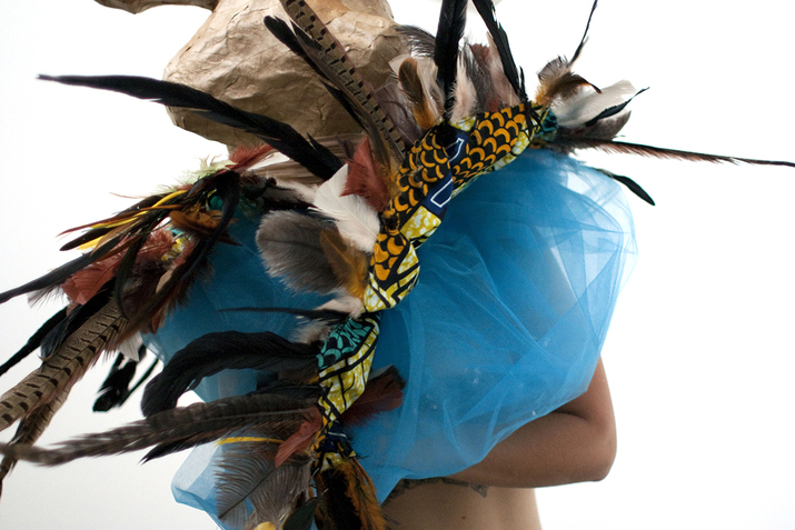 A figure is covered by a mass of ocean blue tulle decorated by mutlicolored feathers that they are holding.