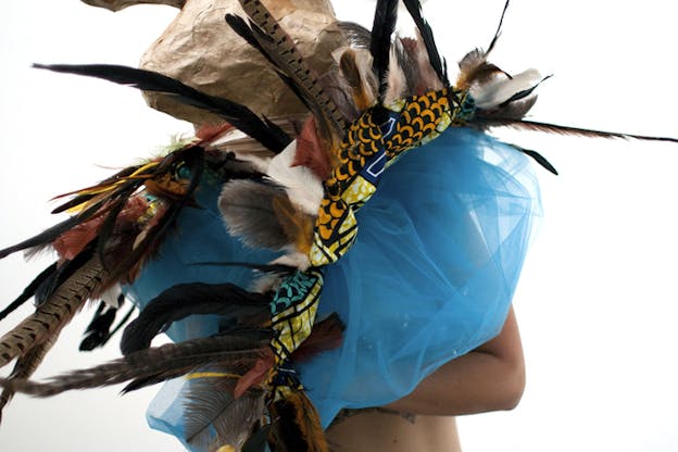 A figure is covered by a mass of ocean blue tulle decorated by mutlicolored feathers that they are holding.