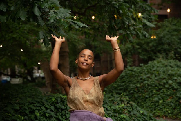 Jasmine Hearn is smiling with their eyes closed, bathed in soft sunlight. Their arms are stretched above their head, elbows slightly bent with their palms facing upward. They wear a necklace, a bracelet on their left arm, and a brown top against a backdrop of trees, bushes, and vines.