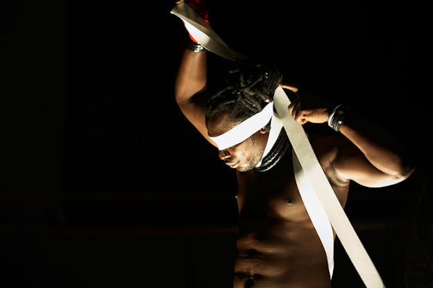 A shirtless performer with ring necklaces adorning their neck covers their eyes with a white ribbon that they pull  upwards from one side.