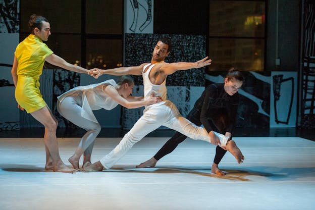 Mitchell wearing white pants and tank top twists his waist looking back at his extended arm as one performer dressed in a black bodysuit holds his extended back leg, one performer dressed in white holds his waist, and a fourth performer dressed in yellow holds his other hand.  