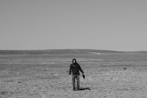 Black and white photograph of Barr standing in the middle of an expansive and remote desert with sparse shrubs and gently sloping hills. 