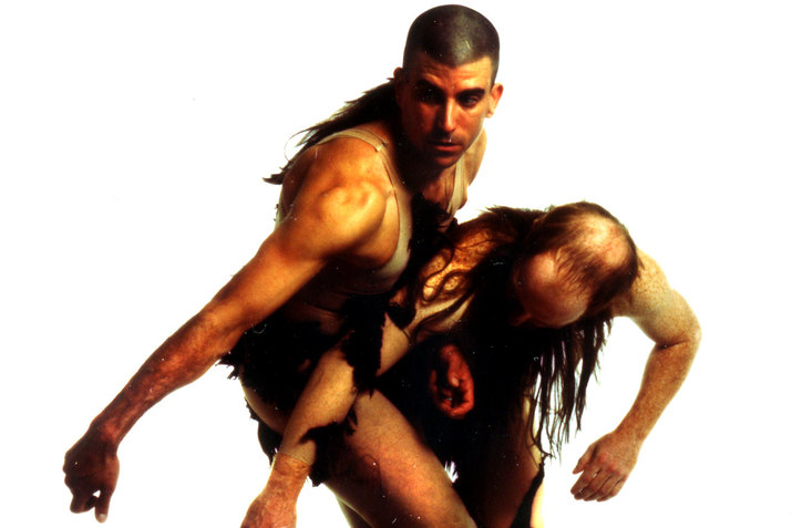 Two muscular persons with long tangled hair wearing minimal clothing twist around each other, one leaning downwards and the other looking to the left in the direction of their extended arm. 