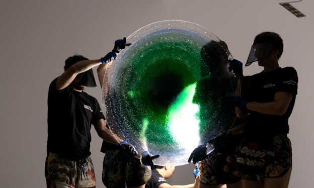 Perfromers wearing face shields hold each side of a circular glass structure. From the center the glass is a deep green color and it becomes lighter further from the center, the outer edges are blue color that turns into transparent.  
