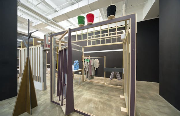 An installation image of a purple painted wooden framed structure besides a white painted wooden structure. On top of the purple structure, there are three oversized spools of thread. Just outside of this structure, there is a small triangular wooden structure. 