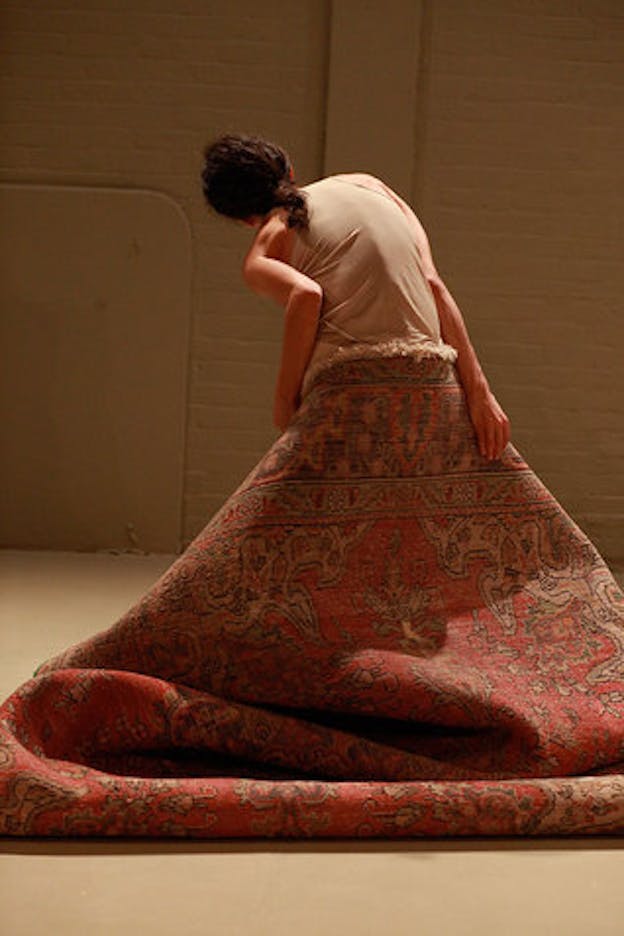 A performance still of Maria Hassabi standing, facing away from the camera, leaning towards the left and dragging a thick red carpet behind her. 