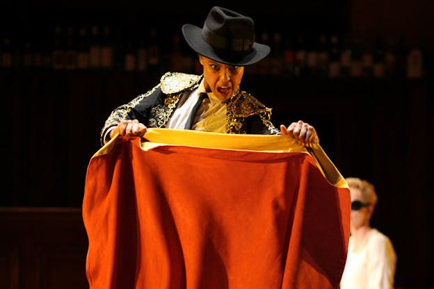 A performance still of a performer dressed in a wide brimmed hat and a bull fighting outfit holding a square of red fabric taught between their two hands. They have an expressive facial expression with their mouth held open as if mid-speech. The background is dark and theres another blurred performer wearing sunglasses on the right.