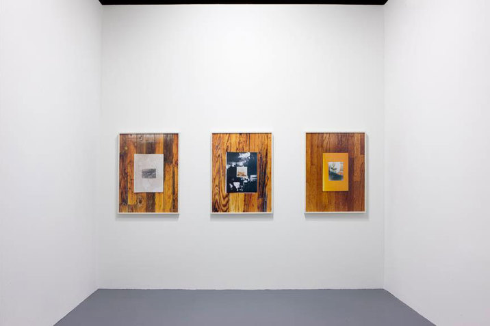 Installation view of three side-by-side framed photographs depicting rectangles of collaged images in the center of wooden floor panels, on a blank white wall. 