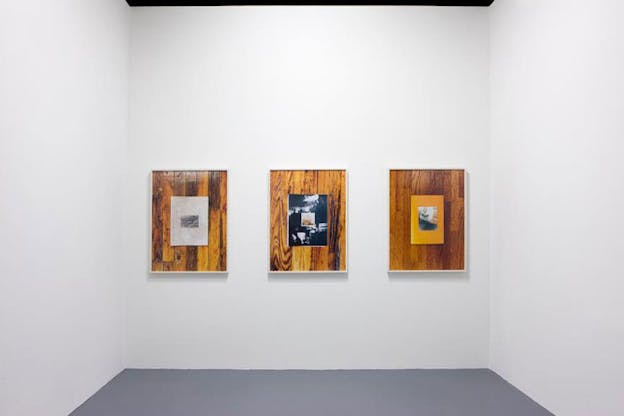 Installation view of three side-by-side framed photographs depicting rectangles of collaged images in the center of wooden floor panels, on a blank white wall. 
