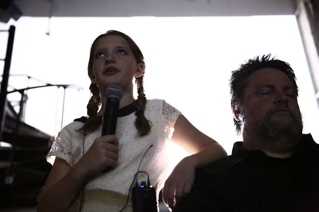 Low-angle shot of young person in two braids and a white-lace blouse looking ahead and speaking into microphone while resting their forearm on Findlay's shoulder, his eyes closed. 