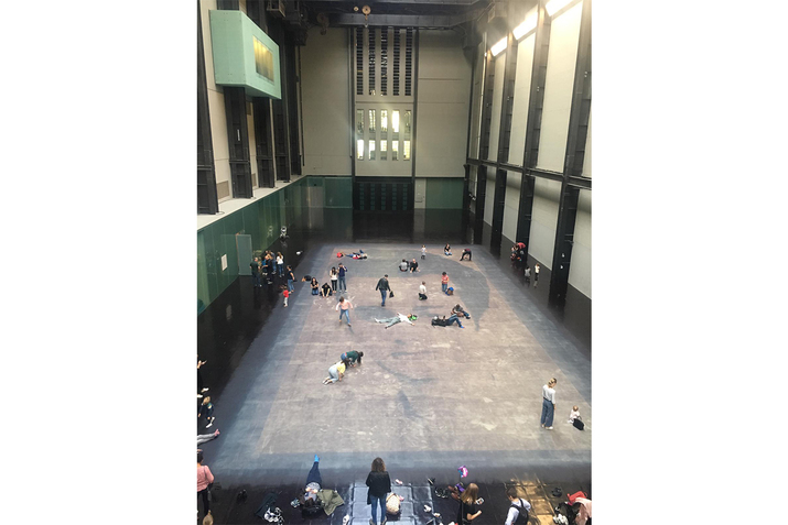An eagle view photograph of people on hall with a high ceiling laying or kneeling on the ground alone and in groups. The floor they are situated on holds the image of a face.