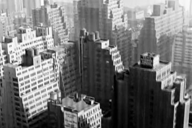 A black and white video still from a bird's eye view overlooking several tall buildings.