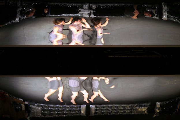 A photograph split horizontally in the middle by a thick black bar. Above the bar, the torsos of three people in purple outfits are seen against a grey background. They lift their left arms above their heads while their right arms are held out at chest height. A distortion filter blurs the middle of their bodies. Below the horizontal bar, the legs of these figures are seen, again distorted by a filter. They step on tip toes. A circular border surrounds the entire photograph and it appears to be constructed from grey pool floats. 
