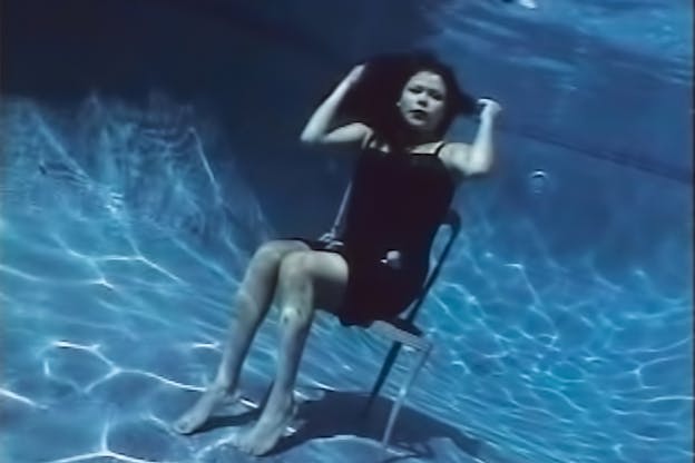 A woman clad in black, sits on a chair underneath the water at the bottom of a pool. The sun illuminates creating thin white lines in the bottom of the pool.