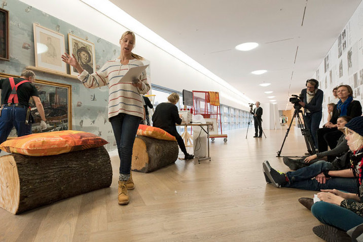 Backstrom raises their right arm while reading from a sheet of paper in a room with two cut logs, two persons filming on a tripod, spectators sit against the right wall, a person sits before a sewing machine, and one person hanging framed paintings. 
